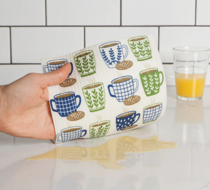 Mano usando una bayeta sueca ecológica con estampado de tazas para limpiar un derrame en la cocina
